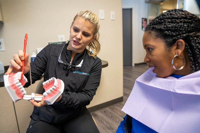 charlotte dental arts hygienist showing african american woman right way to brush using large toothbrush and model of a mouth
