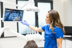 A female dentist in blue scrubs stands in a clinic, pointing at X-rays on a screen, as she explains the dental condition to an attentive patient in a chair.