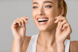 Oral hygiene. Young woman with perfect smile using dental floss, grey studio background