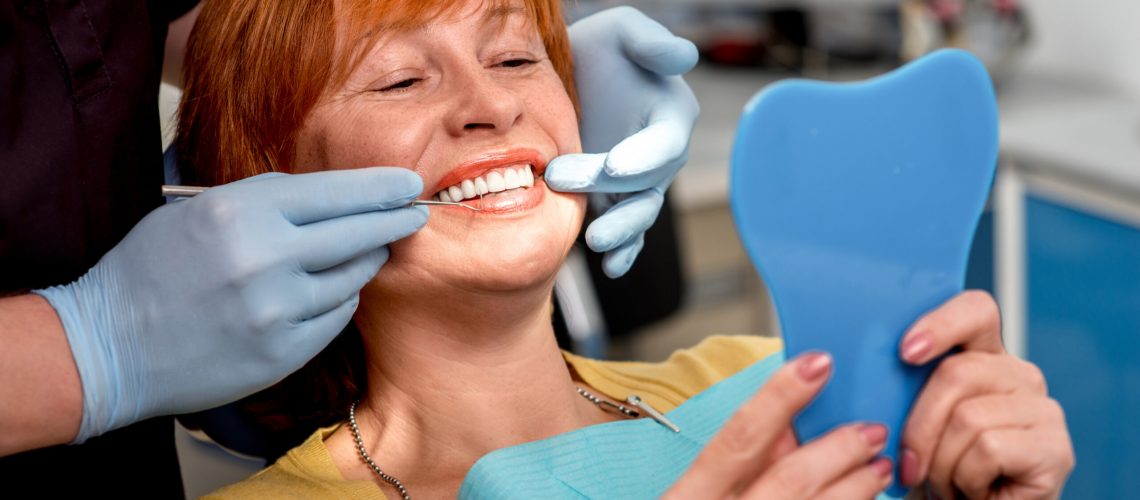Smiling senior woman with new dental implants sitting in the dental office and looking at the mirror.