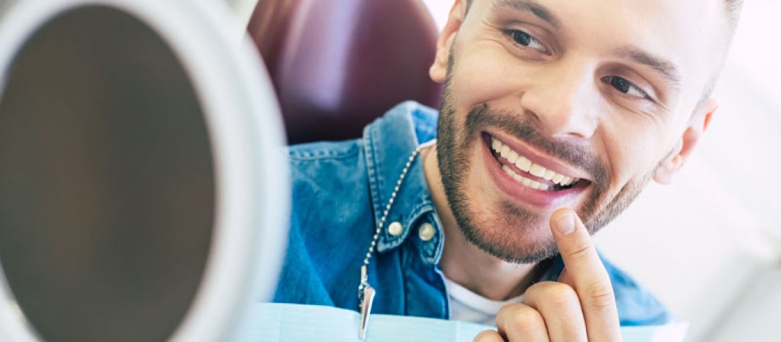 a picture of a dental patient smiling into a hand held mirror and pointing at his new smile because he was just treated with customized dental veneers.